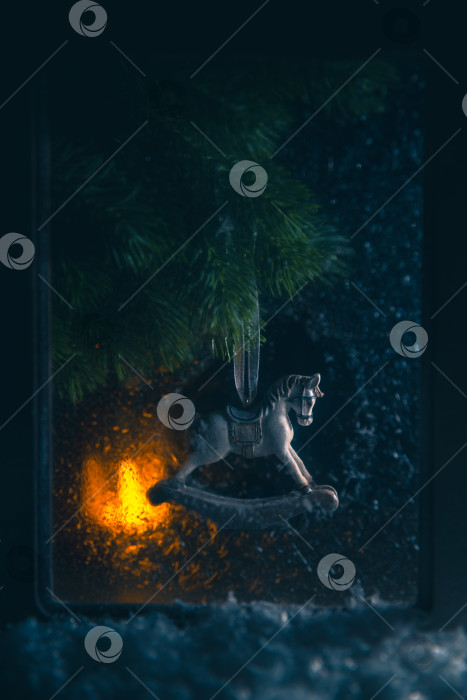 Скачать Украшение в виде лошадки-качалки, висящее на праздничной ветке рождественской елки, создает уютную и ностальгическую атмосферу зимнего праздника, а вид из заснеженного окна создает ощущение тепла и традиций фотосток Ozero