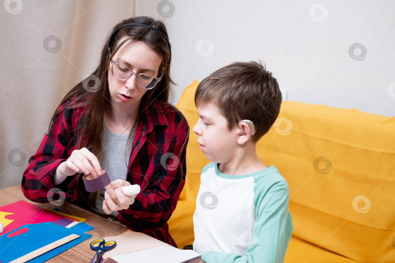 Скачать Молодая женщина в красной клетчатой рубашке и ее сын со слуховыми аппаратами склеивают бумажную гирлянду на Рождество. Рождественский декор для дома своими руками. фотосток Ozero