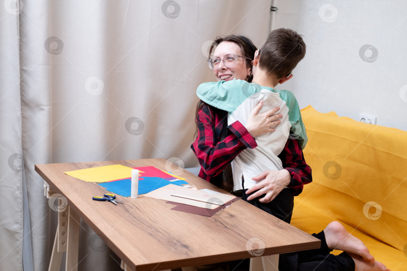 Скачать Молодая женщина в красной клетчатой рубашке с подтяжками обнимает своего сына со слуховыми аппаратами, делая бумажную гирлянду на Рождество. Рождественский декор для дома своими руками. фотосток Ozero