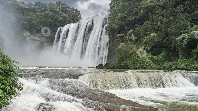 Скачать Живописный тропический водопад. Потоки белой воды стекают со скалы в реку. фотосток Ozero