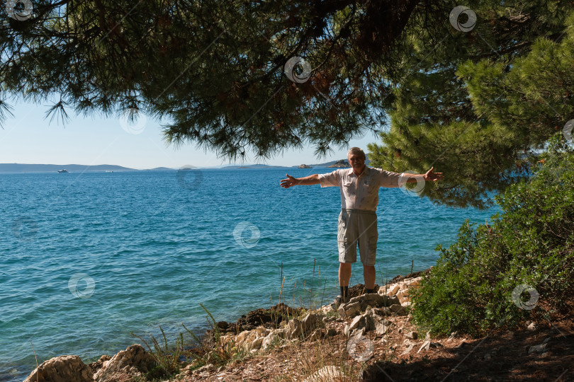 Скачать Счастливый пожилой мужчина раскинул руки на каменистом пляже под ветвями хвойного дерева у моря в Хорватии фотосток Ozero