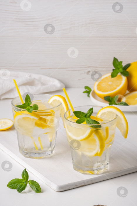 Скачать Освежающие напитки с ломтиками лимона и листьями мяты на белой поверхности в ярком оформлении и свежими фруктами на заднем плане фотосток Ozero