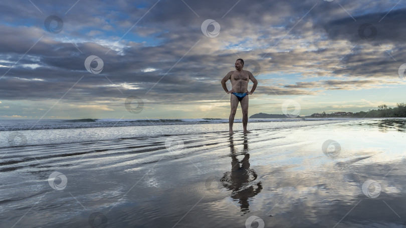 Скачать Рассвет над океаном. Мужчина в плавках стоит на песчаном пляже фотосток Ozero