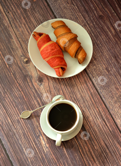 Скачать Чашка черного кофе на блюдце с ложечкой и тарелка с двумя свежими круассанами с шоколадно-малиновой начинкой на деревянном столе. фотосток Ozero