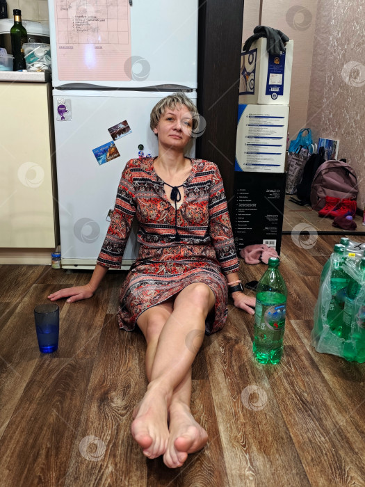 Скачать Уставшая женщина в платье сидит на полу у холодильника с телефоном в руках. Усталая женщина в платье сидит на полу у холодильника. фотосток Ozero