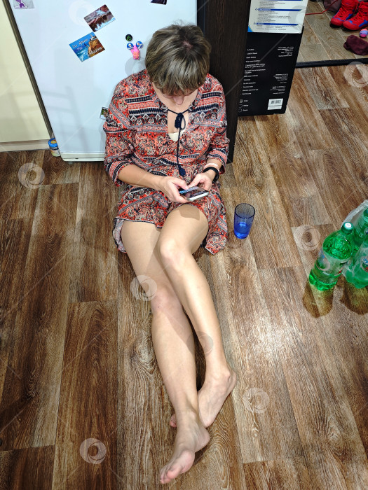 Скачать Уставшая женщина в платье сидит на полу у холодильника с телефоном в руках. Усталая женщина в платье сидит на полу у холодильника. фотосток Ozero