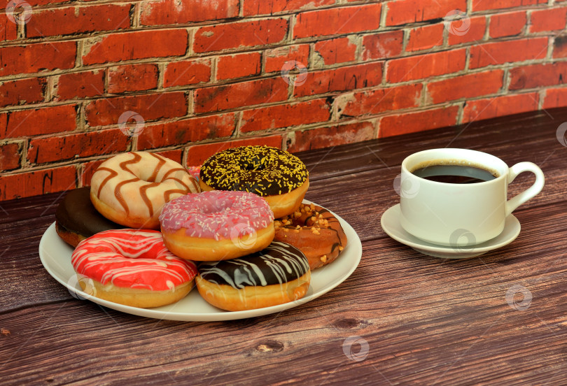 Скачать Чашка черного кофе на блюдце и тарелка с несколькими свежеиспеченными пончиками в разноцветной глазури на деревянном столе. фотосток Ozero