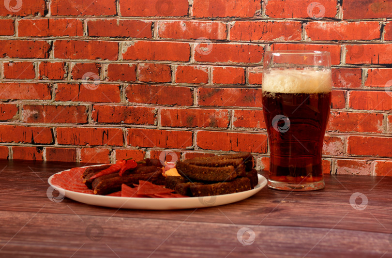 Скачать Высокий бокал полутемного пива и тарелка с разнообразными мясными закусками, гренками и сыром на деревянном столе. фотосток Ozero