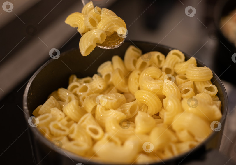 Скачать Макароны готовые, сваренные в черной кастрюле с ручкой. На ложке лежат несколько макаронных трубочек фотосток Ozero