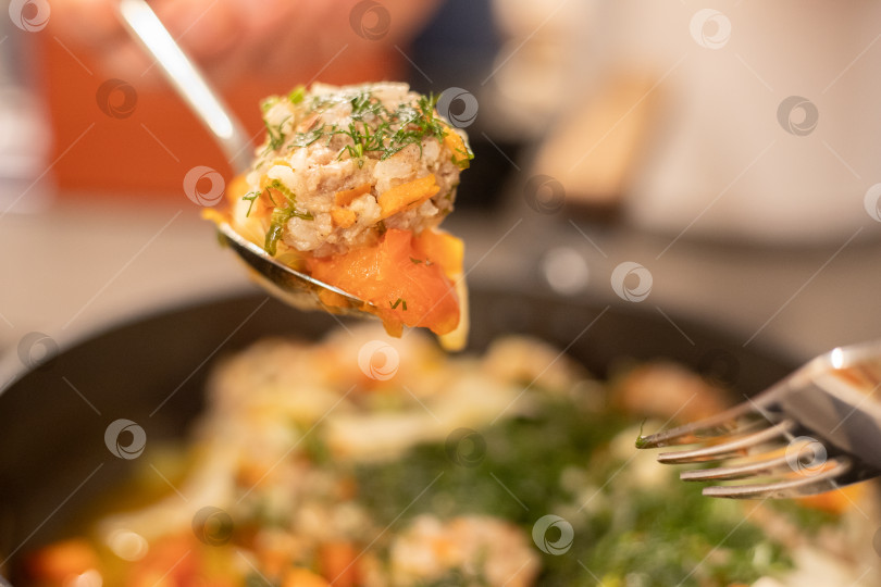 Скачать Готовое блюдо, мясные фрикадельки на пару из фарша с рисом и тушеными овощами в глубокой сковородке, приправленные зеленью и специями. На большой ложке кусок приготовленной еды рядом вилка. Столовые приборы в воздухе над едой. Здоровая пища. фотосток Ozero