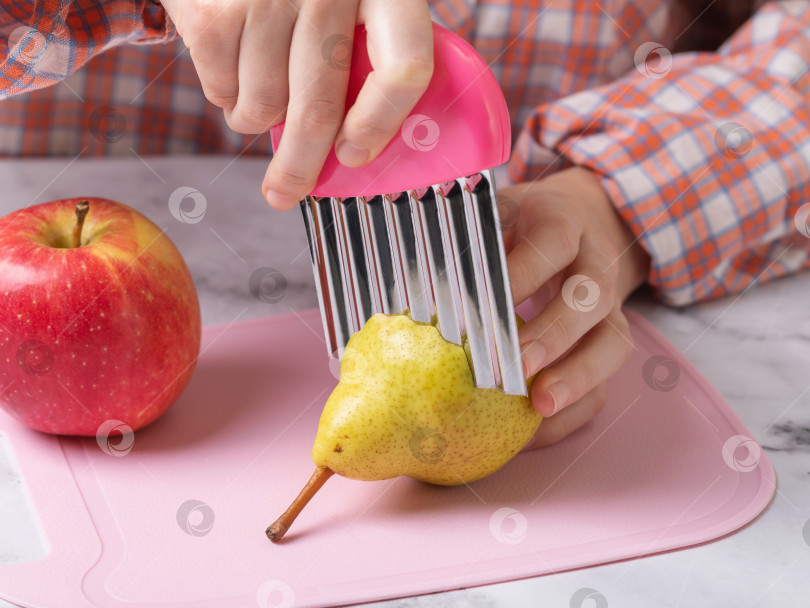 Скачать Ребенок режет грушу волнистым ножом из нержавеющей стали на розовой разделочной доске крупным планом фотосток Ozero