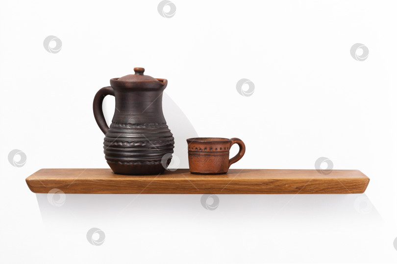 Скачать Глиняный кувшин с ручкой и кружка стоят на дубовой деревянной полке, прикрепленной к белой стене, вид спереди. фотосток Ozero