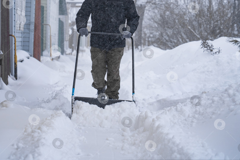Скачать Мужчина в темной куртке разгребает снег лопатой возле дома, деревенская улица, зима, сугробы фотосток Ozero