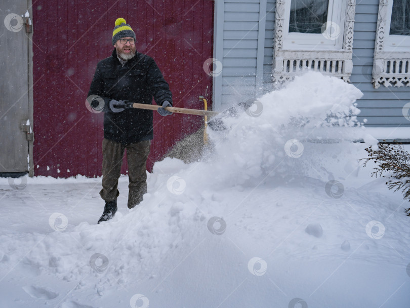Скачать Мужчина в темной куртке разгребает снег лопатой возле дома, деревенская улица, зима, сугробы фотосток Ozero