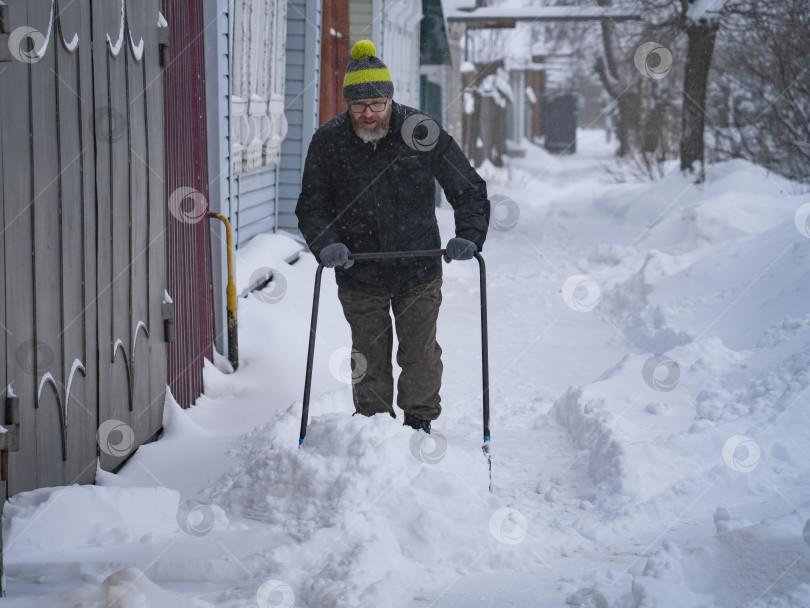 Скачать Мужчина в темной куртке разгребает снег лопатой возле дома, деревенская улица, зима, сугробы фотосток Ozero