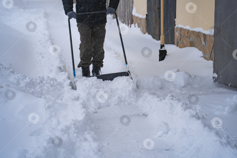 Скачать Мужчина в темной куртке разгребает снег лопатой возле дома, деревенская улица, зима, сугробы фотосток Ozero