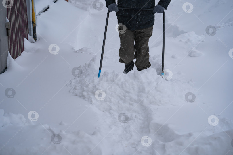 Скачать Мужчина в темной куртке разгребает снег лопатой возле дома, деревенская улица, зима, сугробы фотосток Ozero