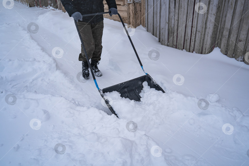 Скачать Мужчина в темной куртке разгребает снег лопатой возле дома, деревенская улица, зима, сугробы фотосток Ozero