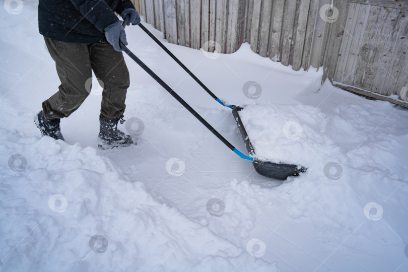 Скачать Мужчина в темной куртке разгребает снег лопатой возле дома, деревенская улица, зима, сугробы фотосток Ozero
