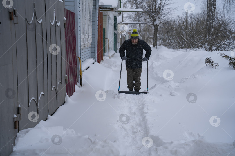 Скачать Мужчина в темной куртке разгребает снег лопатой возле дома, деревенская улица, зима, сугробы фотосток Ozero