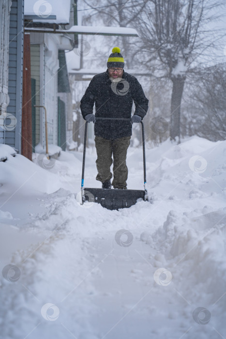 Скачать Мужчина в темной куртке разгребает снег лопатой возле дома, деревенская улица, зима, сугробы фотосток Ozero
