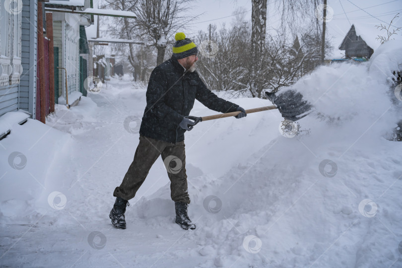Скачать Мужчина в темной куртке разгребает снег лопатой возле дома, деревенская улица, зима, сугробы фотосток Ozero