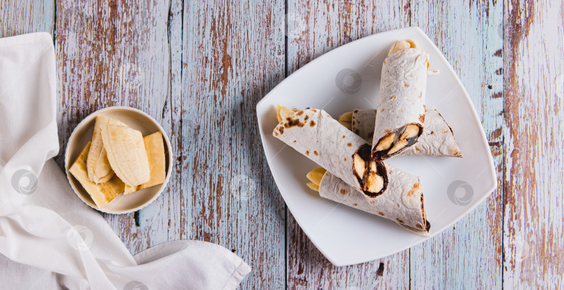 Скачать Покрытые шоколадом ломтики банана, завернутые в тортилью, на тарелке на столешнице посмотреть веб-баннер фотосток Ozero