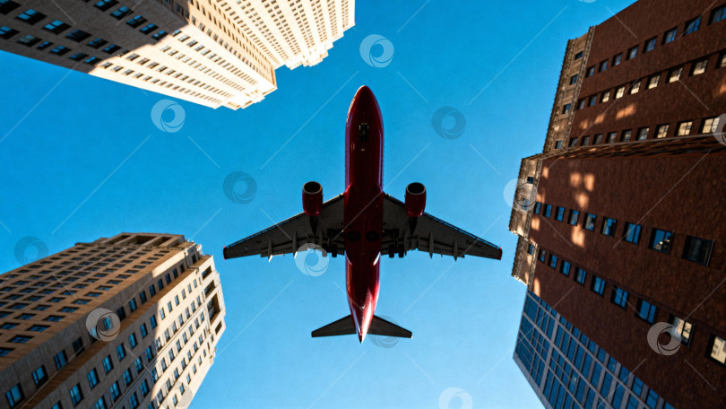 Скачать Фотография самолета, пролетающего между высокими зданиями, снятая с нижнего ракурса на фоне голубого неба. Подходит для тем, связанных с путешествиями, авиацией, городскими пейзажами и современной архитектурой. фотосток Ozero