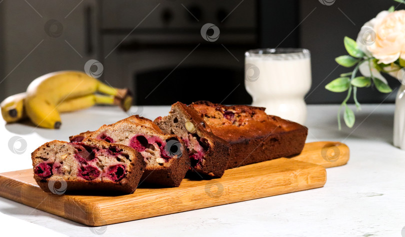 Скачать Банановый хлеб с ягодами, нарезанный ломтиками на деревянной доске. Рядом с ним стоит стакан молока, а на заднем плане изображены бананы и цветы фотосток Ozero