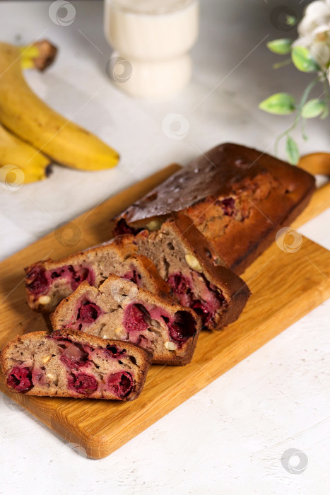 Скачать Банановый хлеб нарезают на разделочной доске. Внутри хлеба видны красные вишни. На заднем плане несколько бананов. Освещение яркое и теплое. Банановый хлеб с ягодами - полезный перекус на завтрак фотосток Ozero