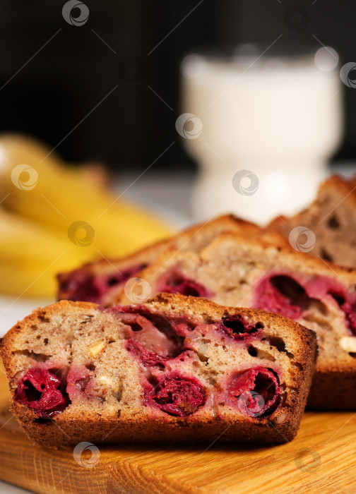 Скачать На деревянной разделочной доске разложены ломтики бананового хлеба с вишней. На заднем плане изображены бананы и стакан молока. Декорация - кухня. фотосток Ozero