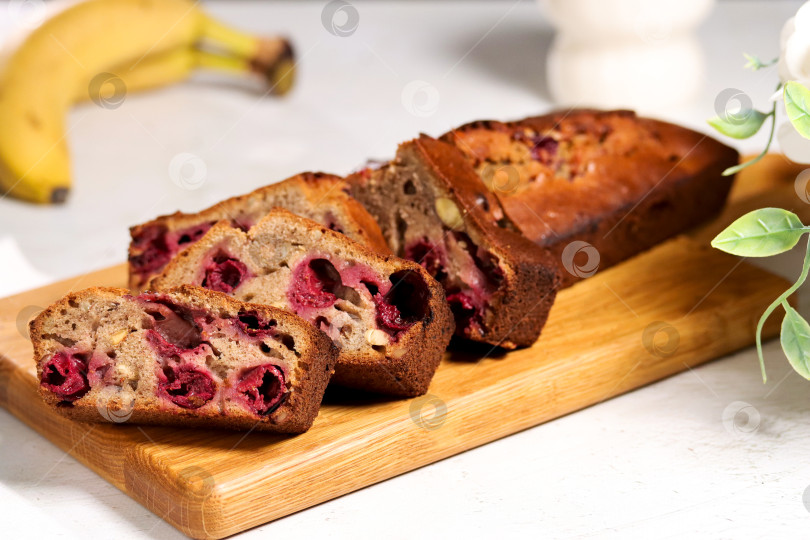 Скачать Свежеиспеченный банановый хлеб с вишней на деревянной доске в яркой кухонной обстановке. Ломтики бананового хлеба с вишней на деревянной разделочной доске. Банан и цветы на утренней сцене фотосток Ozero