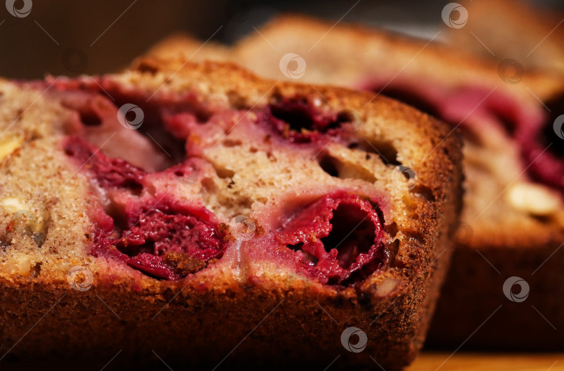 Скачать Банановый хлеб с вишней нарезают на деревянной разделочной доске. Хорошо видна нежная текстура и красные кусочки. На заднем плане - кухонные принадлежности, крупным планом фотосток Ozero