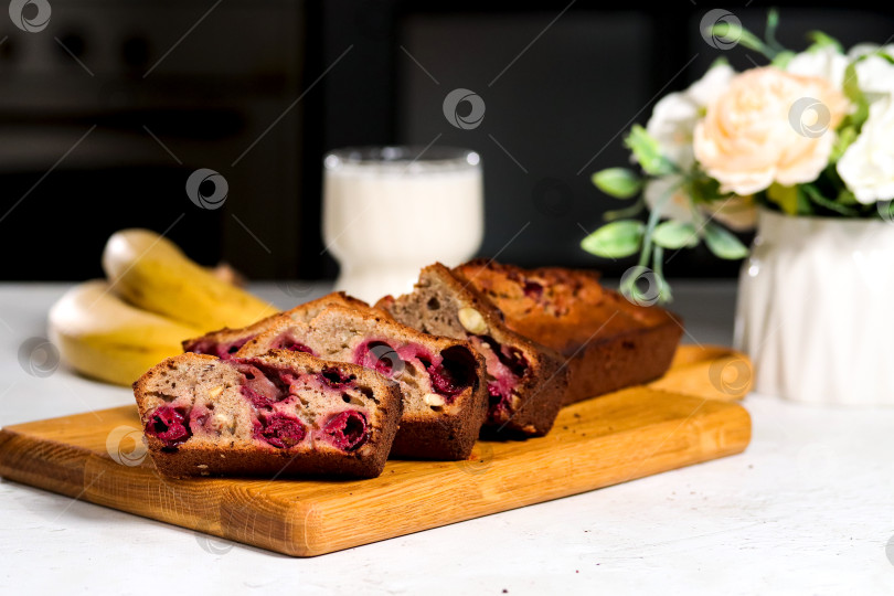 Скачать Банановый хлеб с ягодами на деревянной доске в окружении цветов и стакана молока. Ломтики бананового хлеба с вишней - идеальный вариант для семейного завтрака. фотосток Ozero