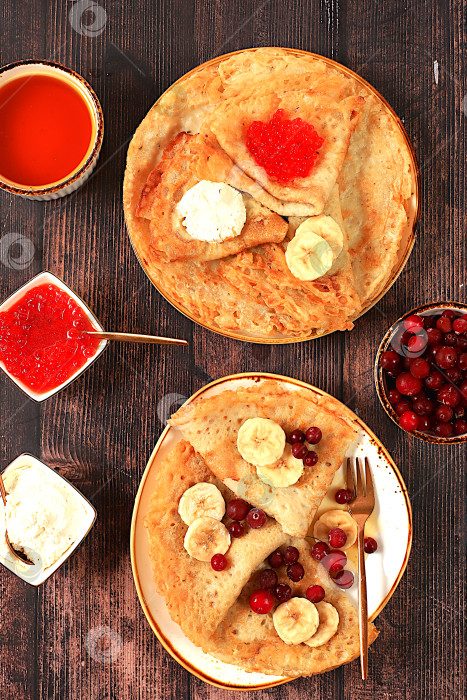 Скачать Вкусные русские блинчики с мягким сыром, бананами, красной икрой и клюквой на кухонном столе. Полезный масленичный завтрак, приготовленный из свежих продуктов. фотосток Ozero