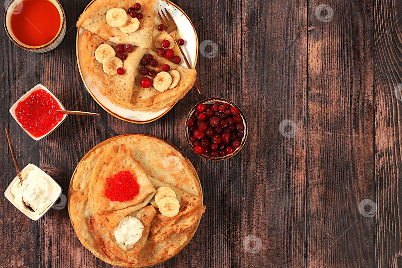 Скачать Вкусные русские блинчики с мягким сыром, бананами, красной икрой и клюквой на кухонном столе. Полезный масленичный завтрак, приготовленный из свежих продуктов. фотосток Ozero
