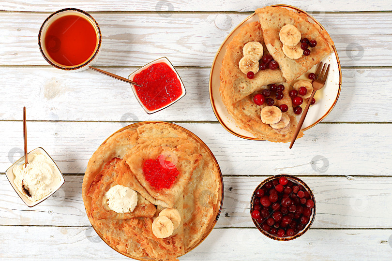 Скачать Вкусные русские блинчики с мягким сыром, бананами, красной икрой и клюквой на кухонном столе. Полезный масленичный завтрак, приготовленный из свежих продуктов. фотосток Ozero