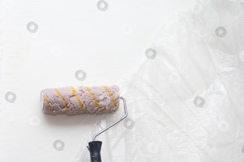 Скачать Покрытый краской валик стоит на фоне белой стены, покрытой защитной пленкой. Ремонт дома или квартиры фотосток Ozero