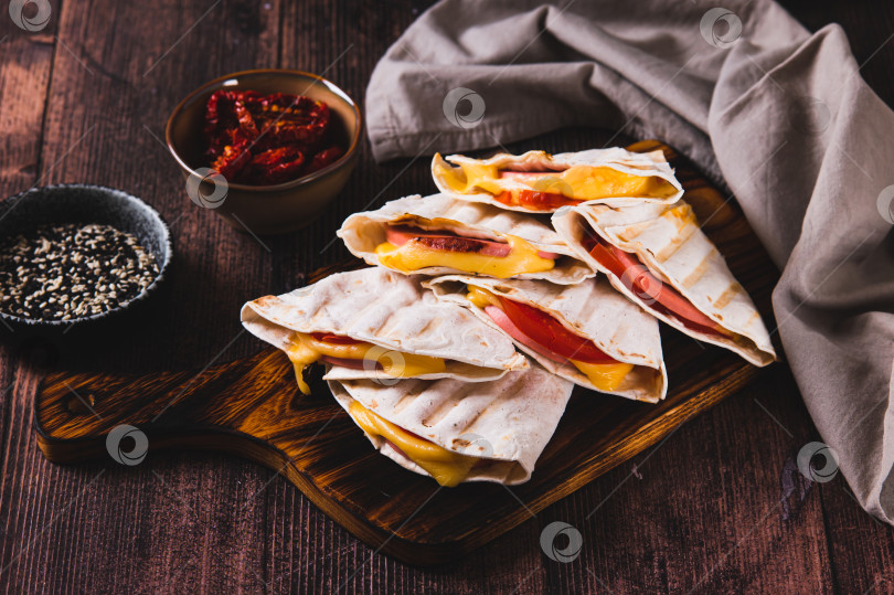 Скачать Колбаса, помидоры и сыр, завернутые в лепешки-гриль, разложенные на доске на столе фотосток Ozero