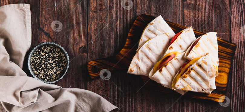 Скачать Веб-баннер с колбасой, помидорами и сыром, завернутыми в лепешки на гриле, выложенный на доске вид сверху фотосток Ozero