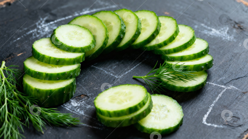 Скачать Свежие дольки зеленого огурца аккуратно нарезанные на темной деревянной доске с зеленью с пробелом для копирования фотосток Ozero