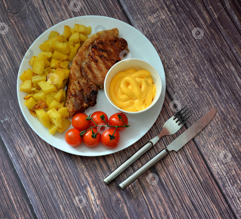 Скачать Антрекот из свинины на гриле с картофелем, помидорами черри и сырным соусом на круглой тарелке на деревянном столе. фотосток Ozero