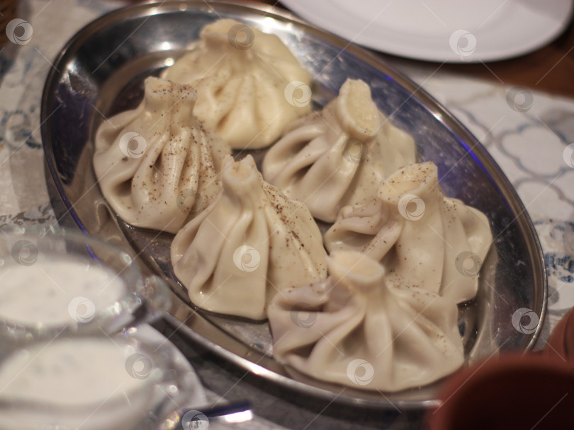 Скачать Традиционные грузинские пельмени хинкали, поданные на металлическом блюде с черным перцем, сметаной и в деревенской посуде. Ресторанная сцена, подходящая для меню, кулинарного блога, путеводителя по еде или статьи о путешествиях. фотосток Ozero