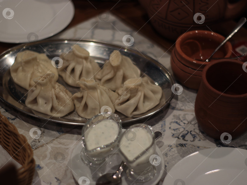 Скачать Традиционные грузинские пельмени хинкали, подаваемые на металлическом блюде с керамическими чашечками и мацони, уютная ресторанная обстановка, подходящая для кулинарного меню, журнала о путешествиях, кулинарной статьи фотосток Ozero