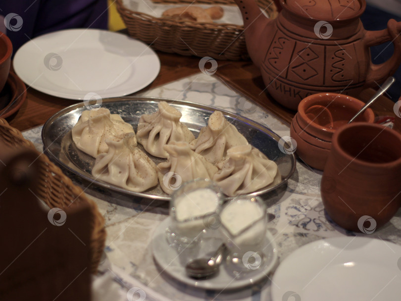 Скачать Традиционные грузинские пельмени хинкали на металлическом блюде с керамическими чашками, мацони и деревенской посудой, уютная ресторанная обстановка, подходящая для кулинарного меню, журнала о путешествиях, кулинарной статьи фотосток Ozero