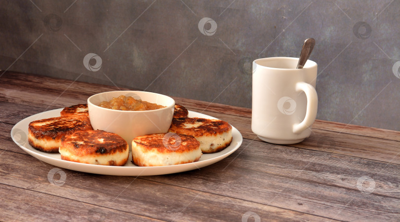 Скачать Свежеприготовленные сырники с изюмом в круглой тарелке, в центре на деревянном столе чашка с яблочным джемом, рядом с ней чашка чая. фотосток Ozero