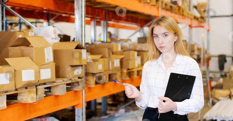 Скачать Женщина-менеджер склада указывает на товар ручкой, держа в руках буфер обмена фотосток Ozero