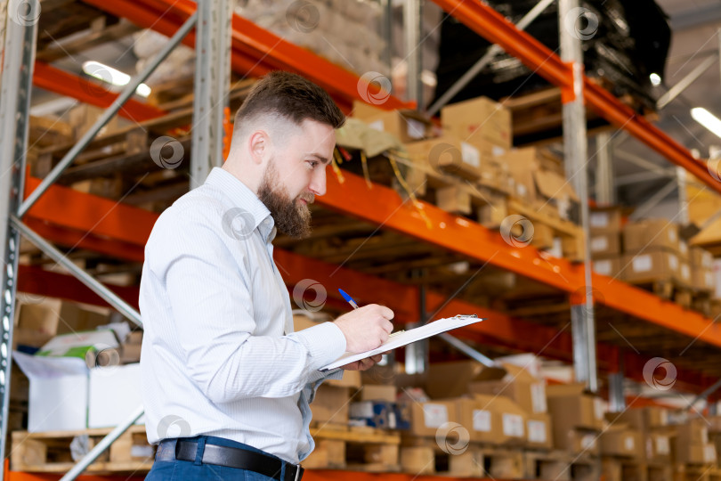 Скачать Бородатый менеджер склада что-то пишет в планшете обмена Возле высоких стеллажей фотосток Ozero