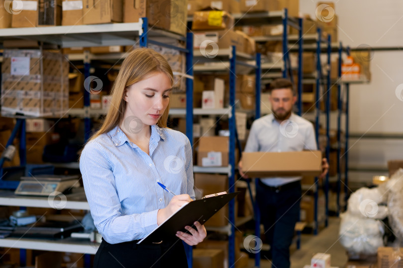 Скачать Сосредоточенная женщина-менеджер по логистике, пишущая в буфер обмена на оживленном складе фотосток Ozero