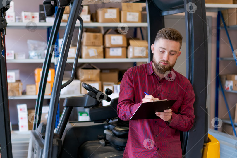 Скачать Сосредоточенный начальник склада что-то пишет в планшете обмена рядом с вилочным погрузчиком фотосток Ozero